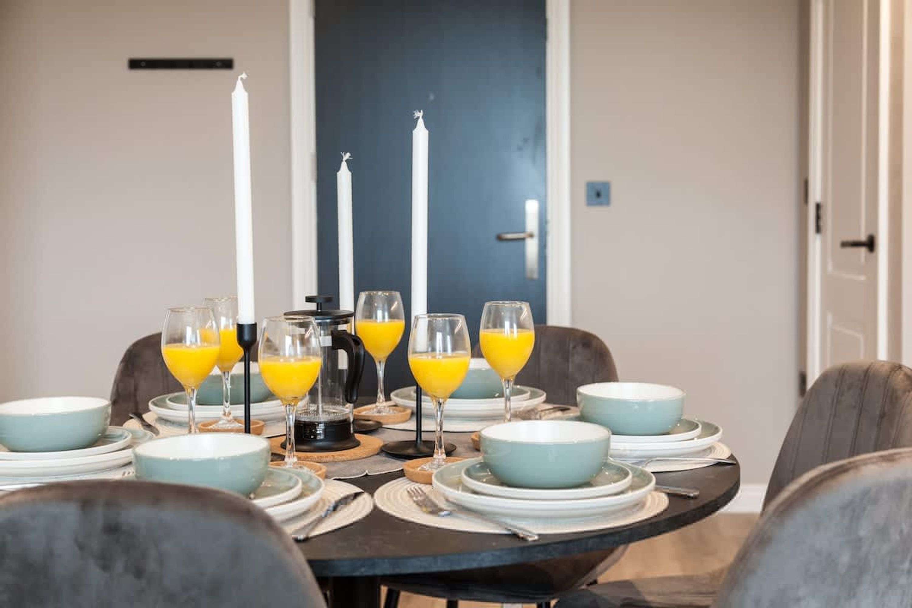 Una mesa de comedor dispuesta con platos, vasos y velas, con una puerta azul al fondo.