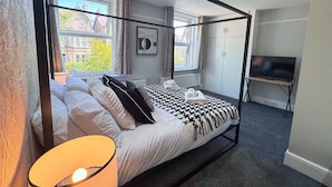 Ein modernes Schlafzimmer mit einem großen Bett, einem Fernseher und einem Fenster mit Blick auf Gebäude.
