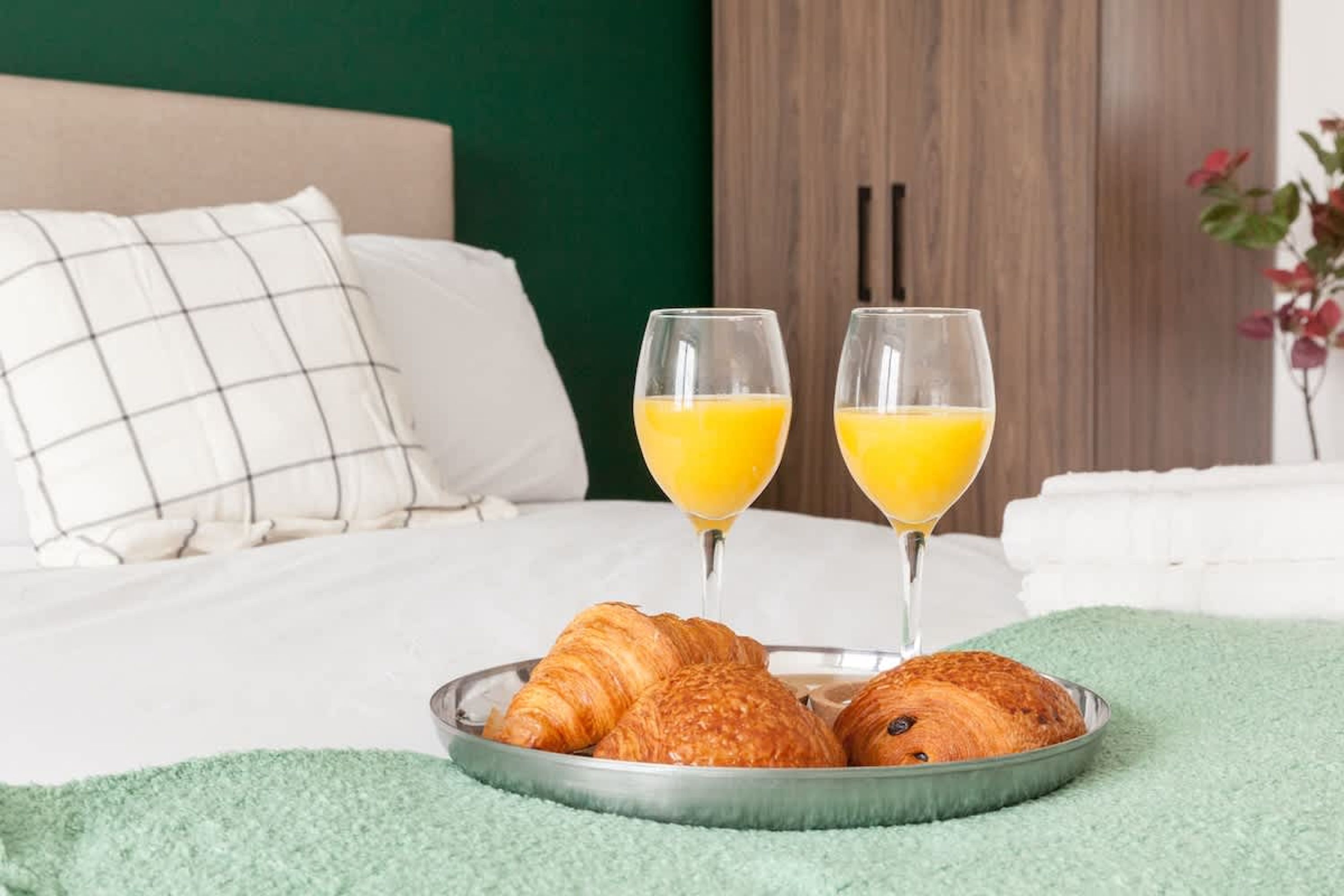 A bed with pillows, a tray with croissants and orange juice glasses, and a green blanket.