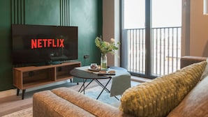 A living room with a large flat-screen TV, a dining table, a sofa, and a balcony with a railing. - 3 Bed Flats Near Station City Centre - Sleeps 6 (Leeds)