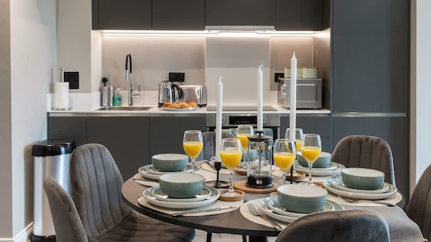 A modern kitchen with dark cabinets, a central island with a coffee machine, and a dining area set for breakfast with plates, glasses, and orange juice. - 3 Bed Flats Near Station City Centre - Sleeps 6 (Leeds)