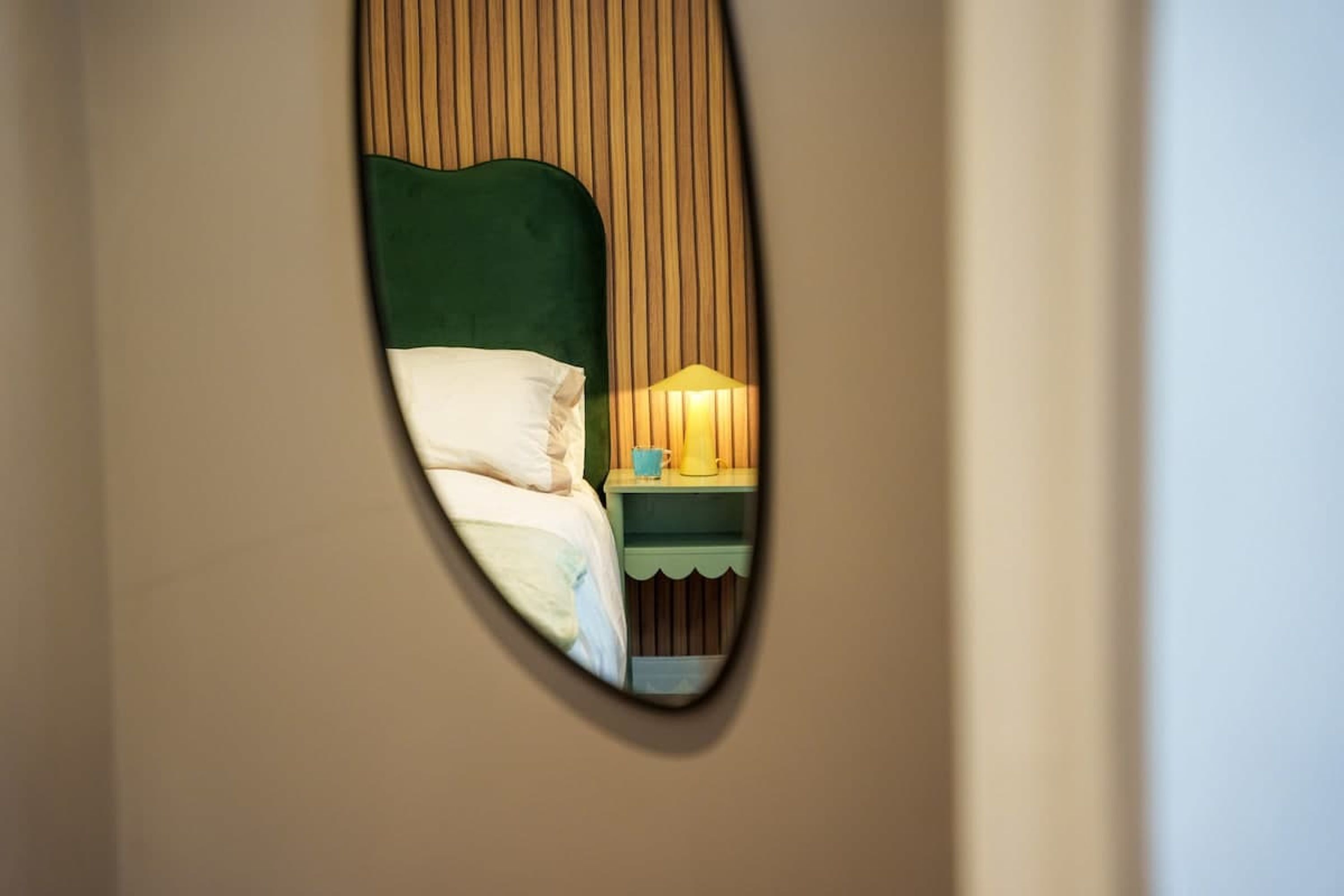 A mirror reflecting a bed with a green headboard, a bedside table with a lamp, and a cup.