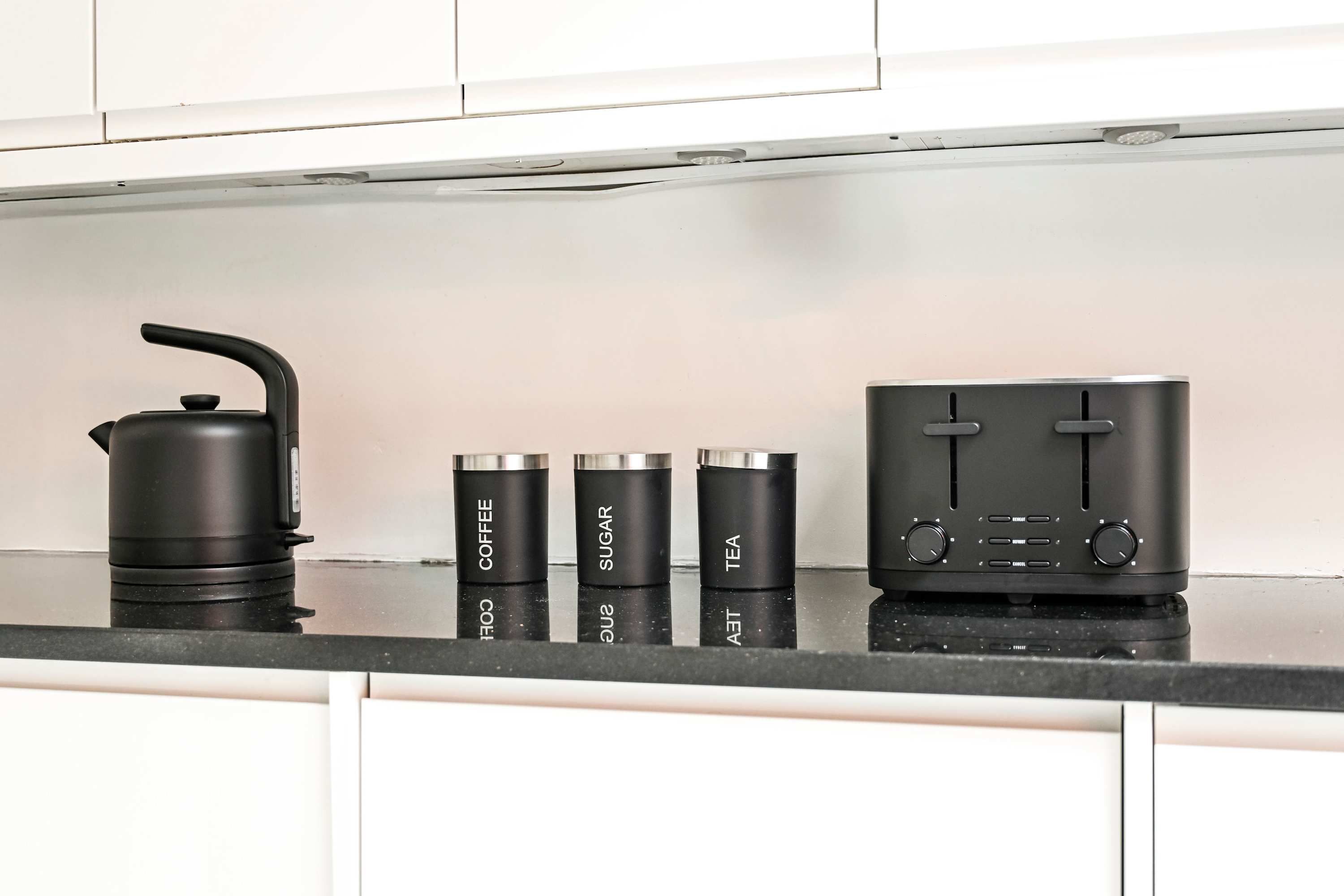 A kitchen counter with a kettle, three spice containers labeled coffee, sugar, and tea, and a black appliance with knobs.