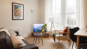 A living room with a sofa, a television, a small table, and a window with curtains. - Bright 2-bed Flat - Harrogate Town Centre - 4 Bed (Harrogate)