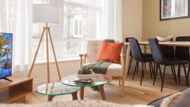 A modern living room with a glass coffee table, a sofa with an orange pillow, a dining area with black chairs, and a lamp with a white shade. - Bright 2-bed Flat - Harrogate Town Centre - 4 Bed (Harrogate)