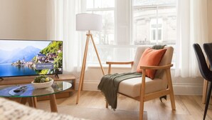 A living room with a television displaying a scenic view, a floor lamp, a chair with a cushion, and a small table with a bowl. - Bright 2-bed Flat - Harrogate Town Centre - 4 Bed (Harrogate)