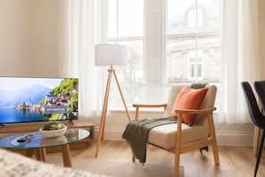 Un salon avec un téléviseur diffusant un paysage, une table basse en verre, un fauteuil avec un coussin, un lampadaire et une vue sur un bâtiment à travers de grandes fenêtres.