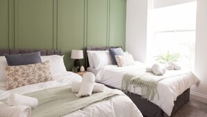 A hotel room with two beds, a green accent wall, a window with a plant, and neatly folded towels. - Bright 2-bed Flat - Harrogate Town Centre - 4 Bed (Harrogate)