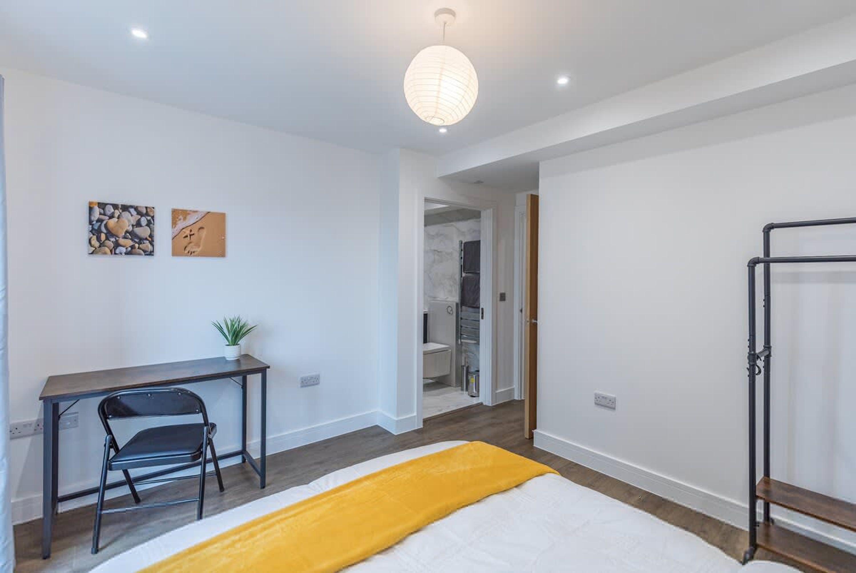 A bedroom with a bed, a desk, a chair, a hanging light fixture, and a shower area.