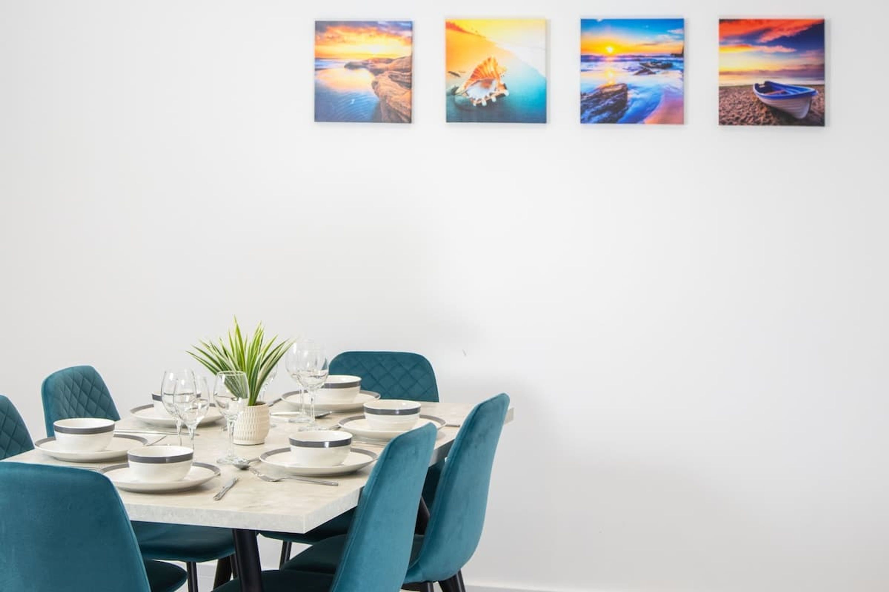 A dining area with a table set for six, teal chairs, and four framed coastal sunset paintings on the wall.