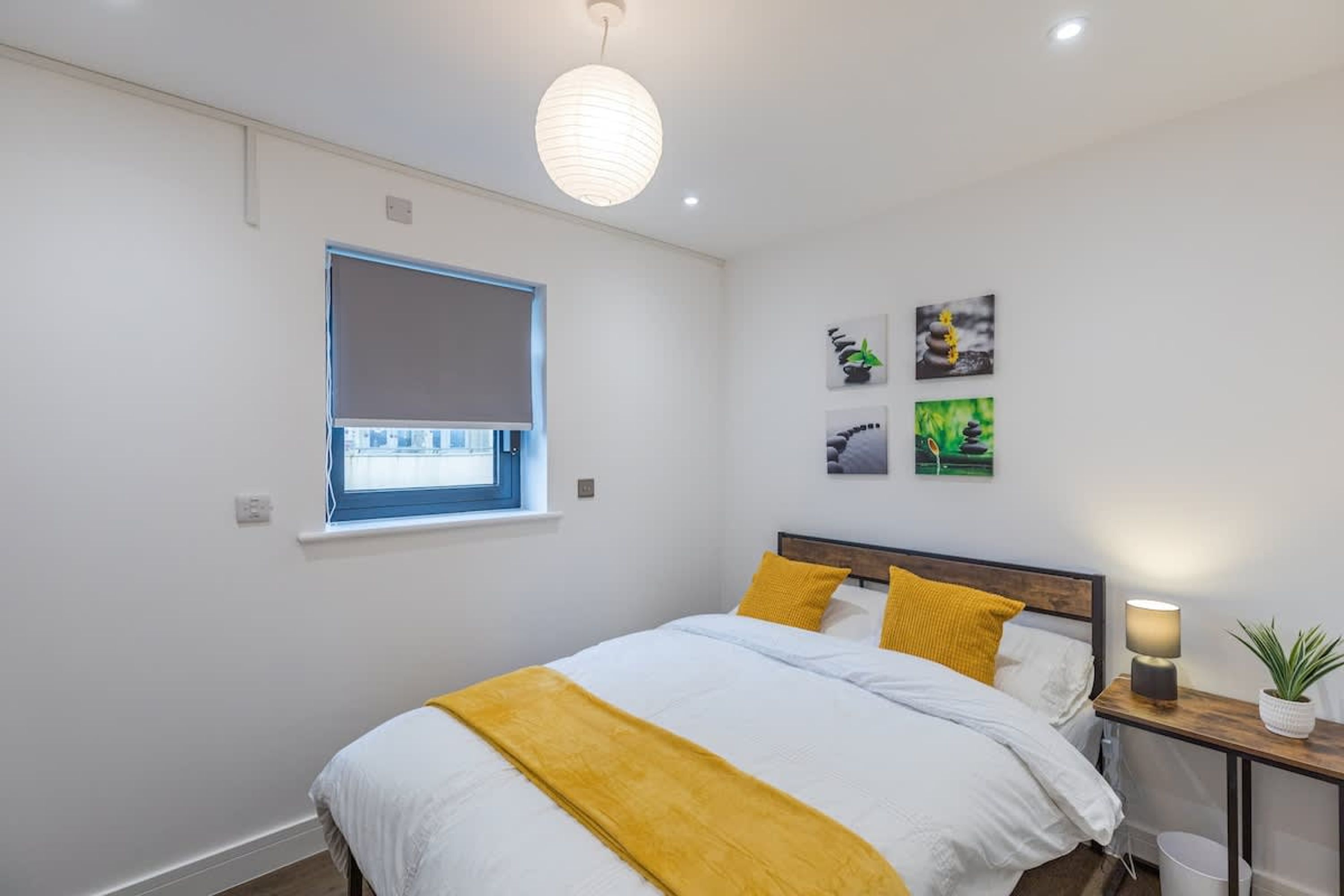Un dormitorio con cama, mesita de noche con lámpara, ventana con persianas y decoración en la pared.