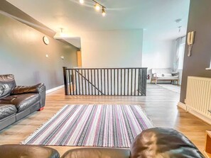 A modern living room with a leather sofa, a patterned rug, a staircase with a railing, and a white chair in the background. - Private 2-bed Duplex Near Headingly w Parking (Leeds)