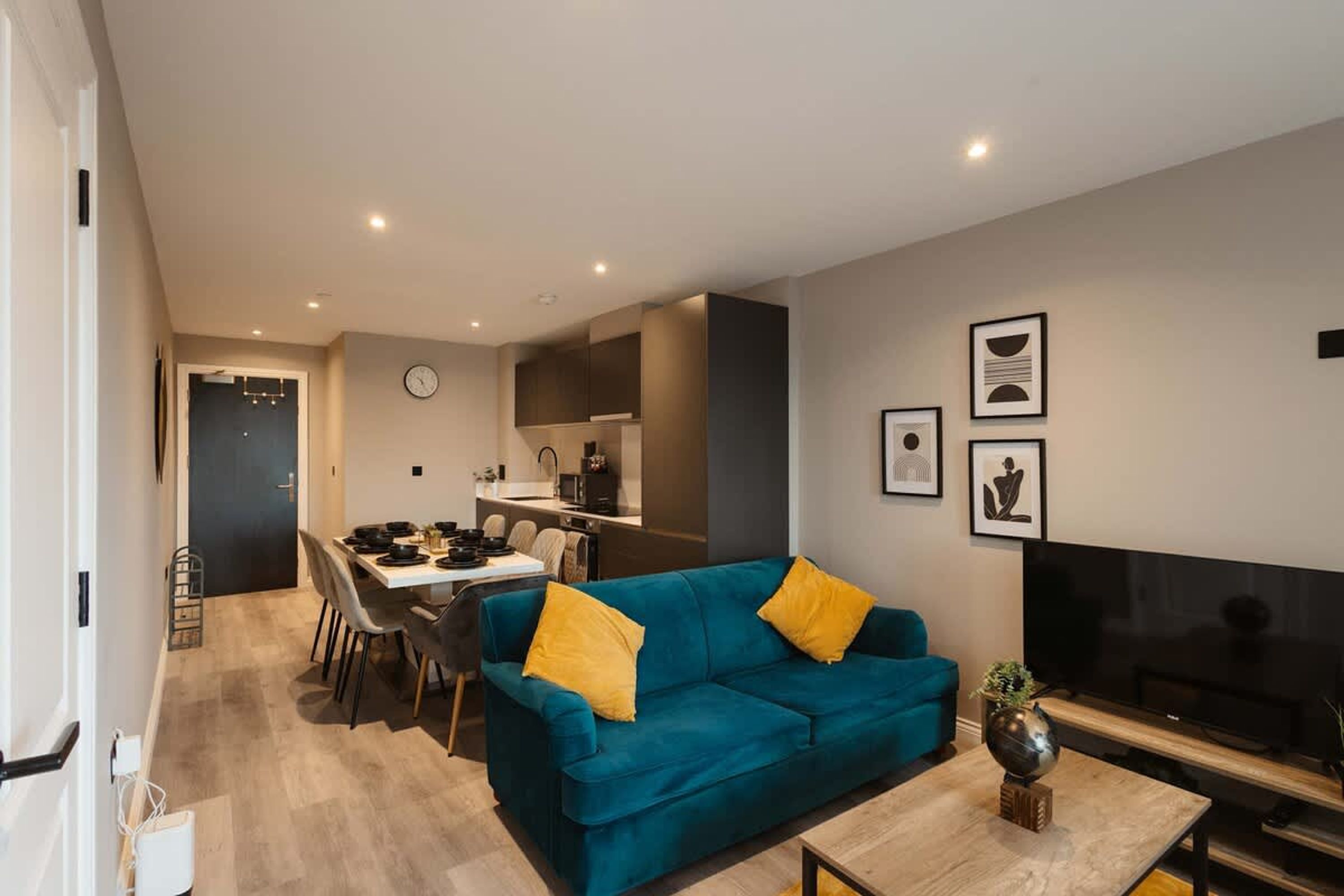 A modern living room with a teal sofa, a dining area, and a kitchen in the background.
