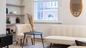 A room with a sofa, a small table with a vase, a black cabinet, and a window with a view of buildings. - Central Leeds Apartment by Station - Sleeps 7 (Leeds)