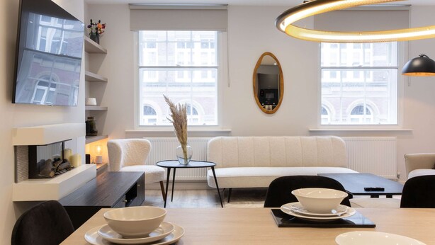 A modern living room with a sofa, dining table, and chairs, a flat-screen TV, and a large window with a view of buildings. - Central Leeds Apartment by Station - Sleeps 7 (Leeds)