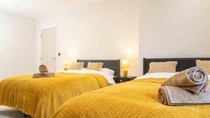 A hotel room with two beds, each with a yellow bedspread and matching pillows, a nightstand with a lamp, and a door. - Central Leeds Apartment by Station - Sleeps 7 (Leeds)