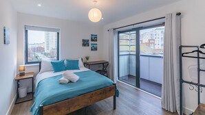 A bedroom with a bed, a desk, a chair, a hanging rack, and a view of the city through a large window. - Seafront Penthouse w/ Balcony Central Brighton (Brighton)