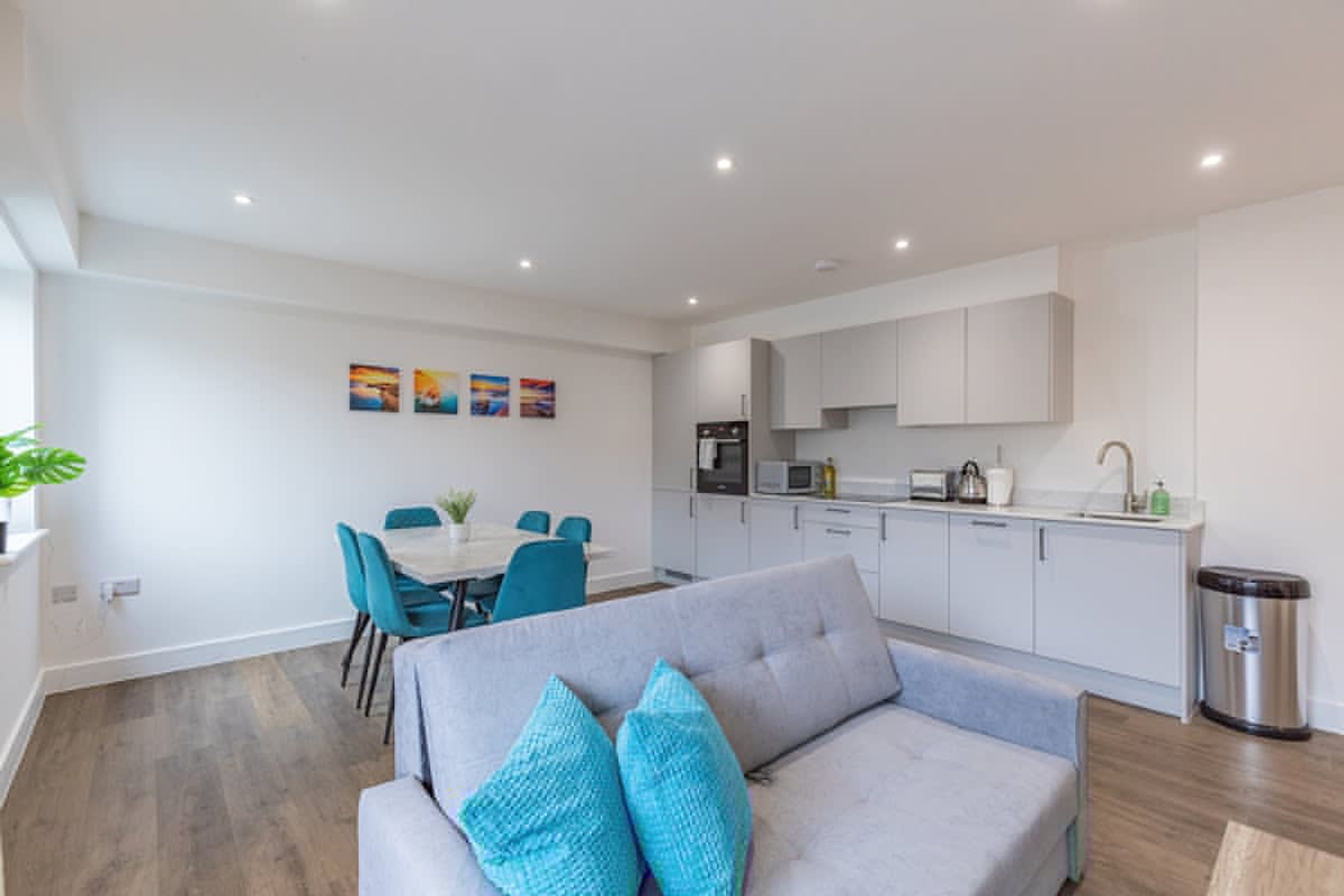 A modern living room with a grey sofa, teal chairs, a dining table, and a kitchen area with white cabinets and appliances.
