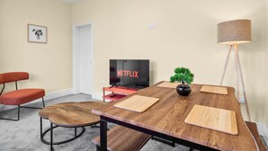 A room with a wooden table, a television, a red chair, a small plant, and a floor lamp. - Modern 3BR Manchester Stay With Parking Wifi (Manchester)