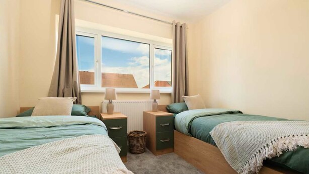 A bedroom with two beds, a window with curtains, bedside tables, and a view of buildings. - Modern 3BR Manchester Stay With Parking Wifi (Manchester)