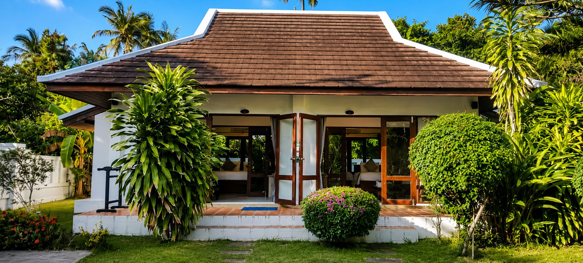 Une maison avec un toit en tuiles, un porche avec un banc et un petit jardin où poussent différentes espèces de plantes.