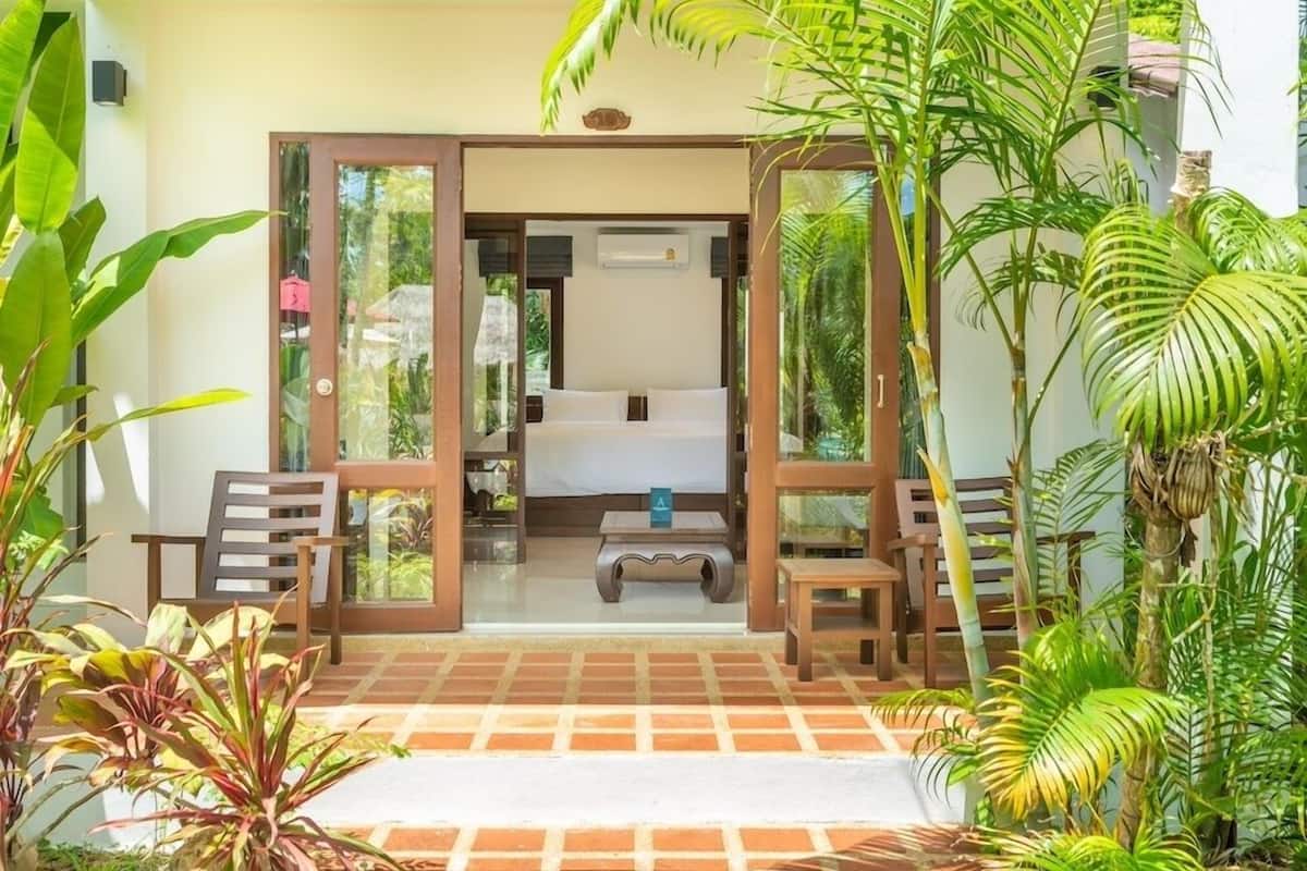 A room with a bed, a small table, and a chair, surrounded by plants and a tiled floor.