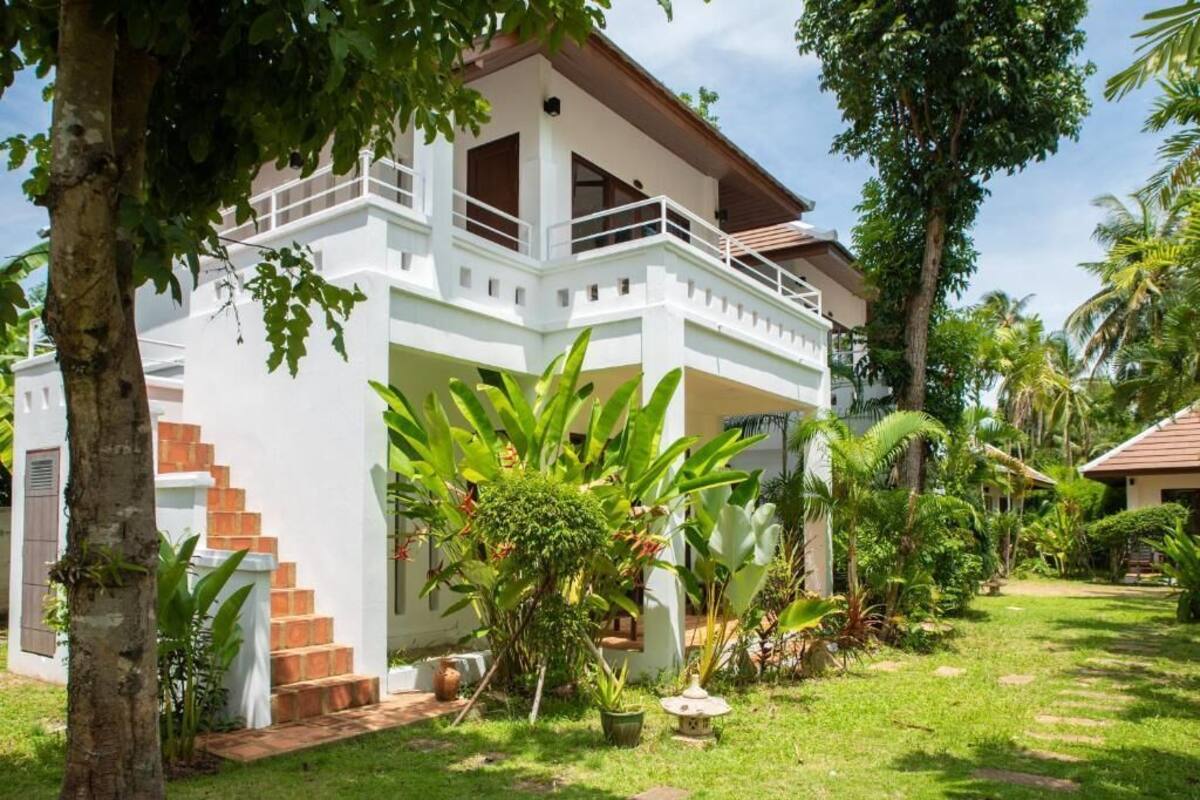 A white two-story house with a balcony, surrounded by lush greenery and palm trees.