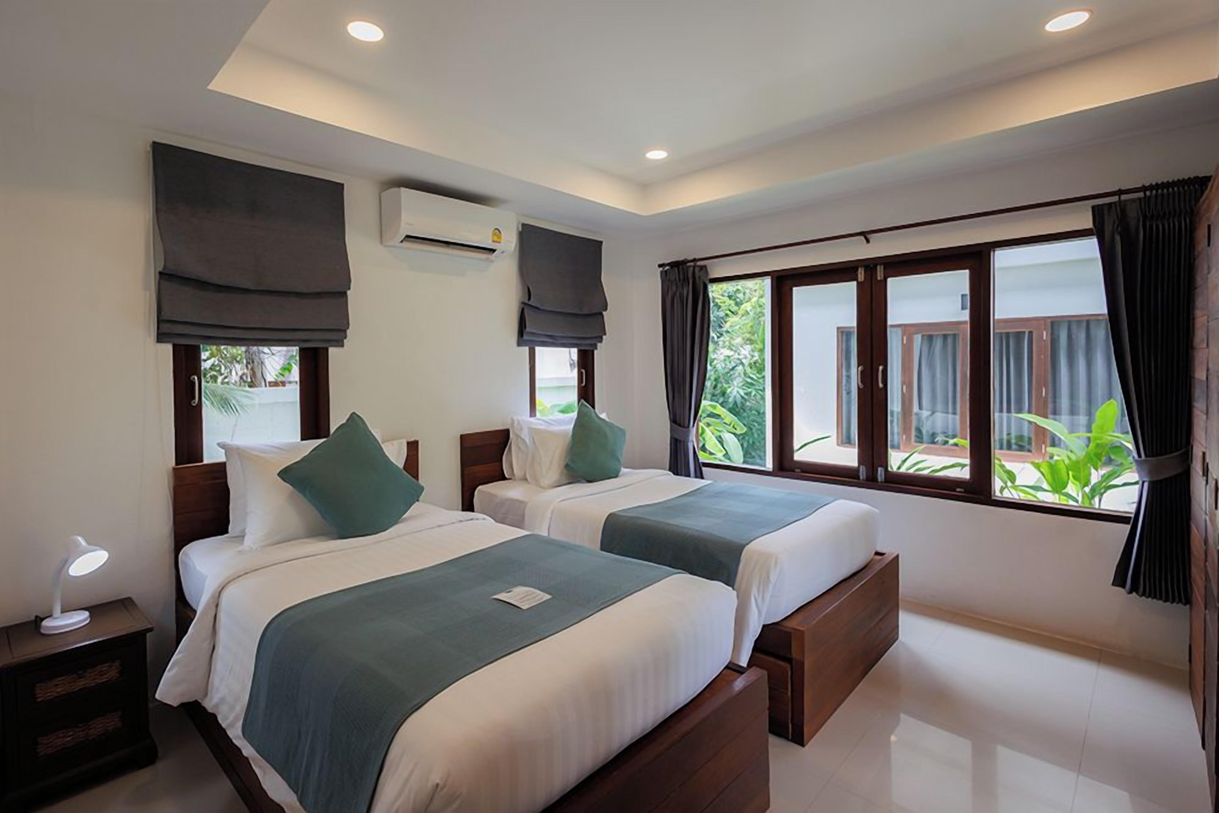 A hotel room with two beds, a nightstand, an air conditioning unit, and large windows with curtains.