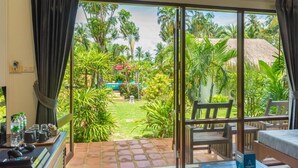 A room with a large sliding glass door leading to a garden with palm trees and a thatched-roof structure. - EASY TIME VILLA by MARALEINA (Koh Samui)