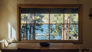 A dining area with a large window, wooden table, and chairs. - Aurora Hygge - Lakefront Luxury Home with Sauna, Hot Tub, Lake Views (Big Bear Lake)