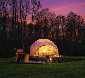 Une structure en forme de dôme géodésique, dotée d’un foyer et d’un espace pour s’asseoir, située dans un cadre boisé.