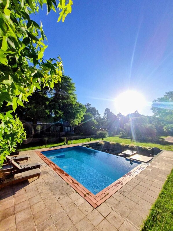 A rectangular swimming pool with a tiled deck, surrounded by greenery and a clear sky.