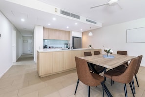 A modern kitchen with a central island, a dining area with a wooden table and brown chairs, and a ceiling fan. - Waterfront Palms Suite: 3 Bedroom apartment in convenient central location (Darwin)