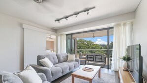 A modern living room with a grey sofa, a wooden coffee table, and a TV on a wooden stand. - Waterfront Lagoon Suite: 1 Bed Apt (Darwin)