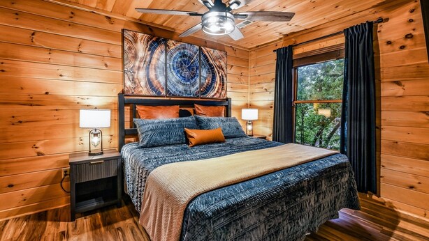 A wooden cabin bedroom with a bed, a nightstand, a ceiling fan, and a window with curtains. - Silver Maple Summit 2 Bedroom Cabin (Gatlinburg)