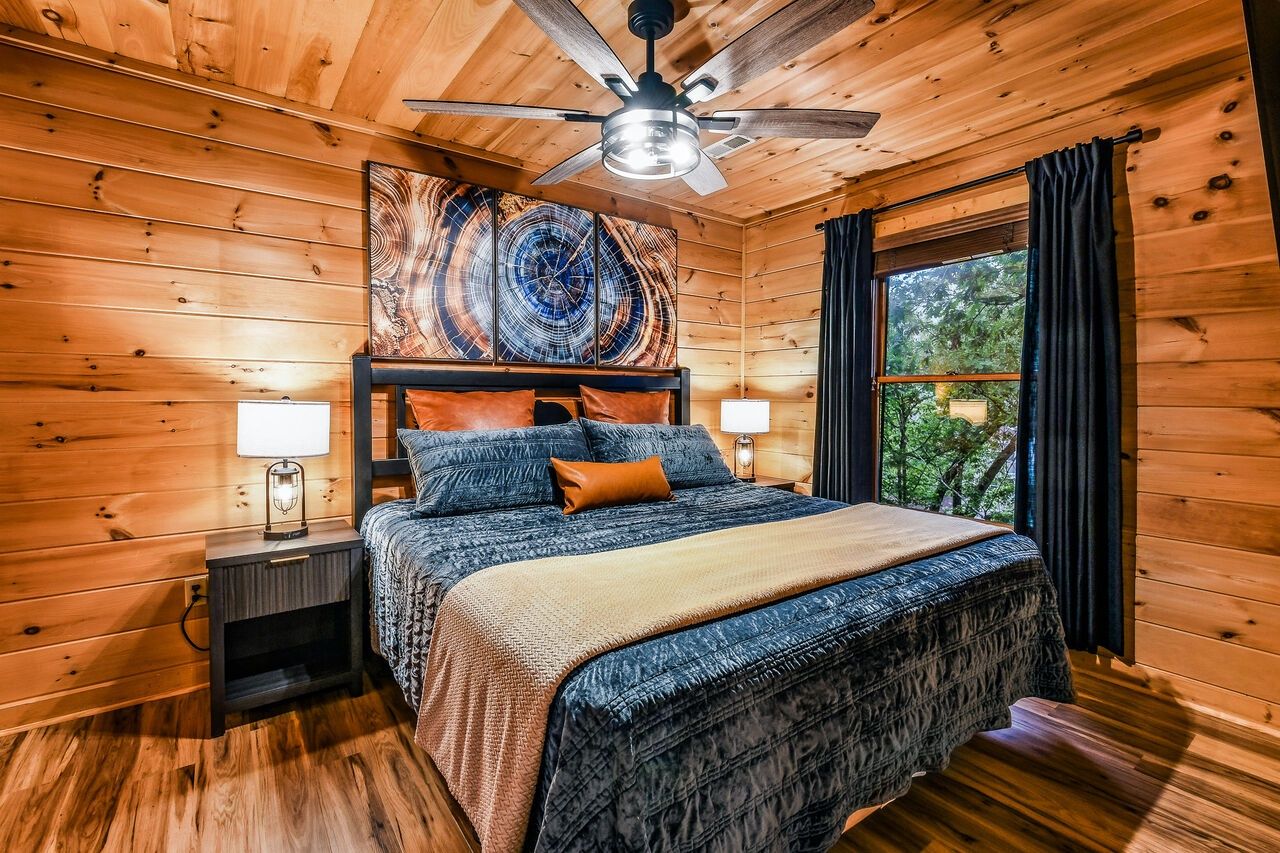 A wooden cabin bedroom with a bed, nightstands, a ceiling fan, and a large window.