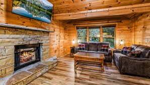 A log cabin interior with a stone fireplace, wooden walls, a large flat-screen TV, a leather sofa, and a wooden coffee table. - Silver Maple Summit 2 Bedroom Cabin (Gatlinburg)