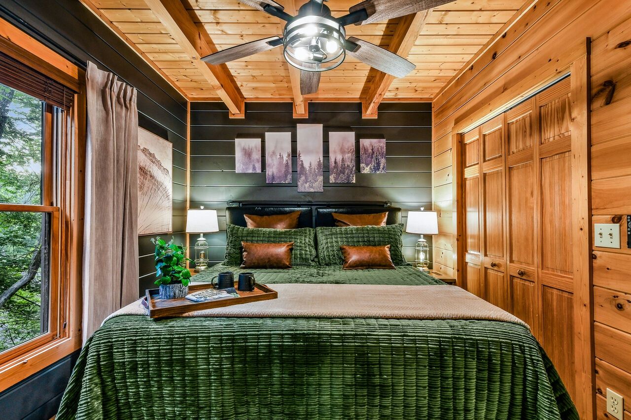 A bedroom with a wooden ceiling, a bed with a green bedspread, a headboard with lamps, and a window with curtains.