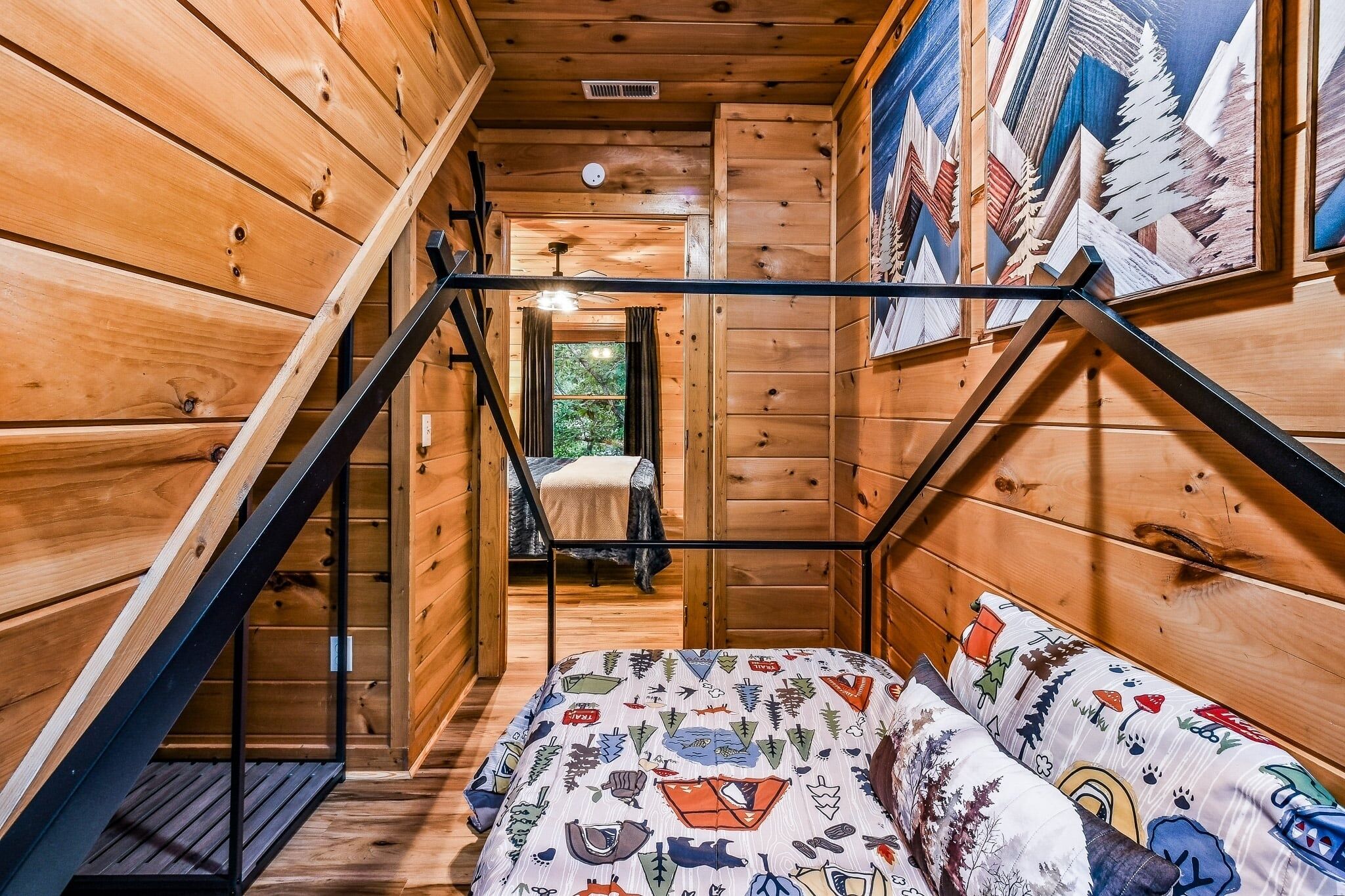 A wooden cabin interior with a bed, a staircase, and a painting of mountains.