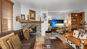 A cozy living room with a stone fireplace, leather sofas, a television, and a bookshelf. - Village Inn Plaza 218 2 Bedroom Condo (Vail)