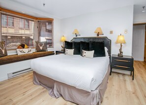 Een slaapkamer met een groot bed, twee nachtkastjes met lampen, een raam met zonwering, en een ingebouwde bank met decoratieve kussens.