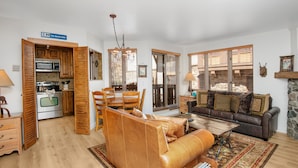 A spacious living area with a leather sofa, a dining table with chairs, a kitchen with wooden cabinets, and large windows with blinds. - Village Inn Plaza 218 2 Bedroom Condo (Vail)