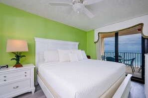 A hotel room with a bed, a nightstand, a lamp, a balcony with chairs, and a view of the ocean. - Phoenix East 804 2 Bedroom Condo (Orange Beach)