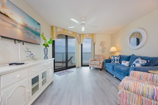A living room with a large TV, a blue sofa, a white cabinet, and a balcony with a view. - Phoenix East 804 2 Bedroom Condo (Orange Beach)