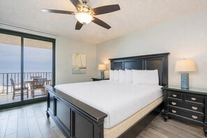 Une chambre d’hôtel avec un grand lit, un ventilateur de plafond, une table de chevet avec une lampe et une vue sur l’océan par une porte-fenêtre coulissante.