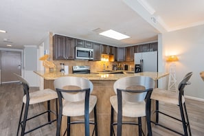 A modern kitchen with dark wood cabinets, a central island with bar stools, and a refrigerator with 'LAUGH' on it. - Phoenix X 514 3 Bedroom Condo (Orange Beach)