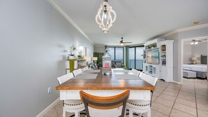 A dining area with a wooden table, white chairs, a chandelier, and a view of the ocean through large windows. - Phoenix X 1504 3 Bedroom Condo (Orange Beach)