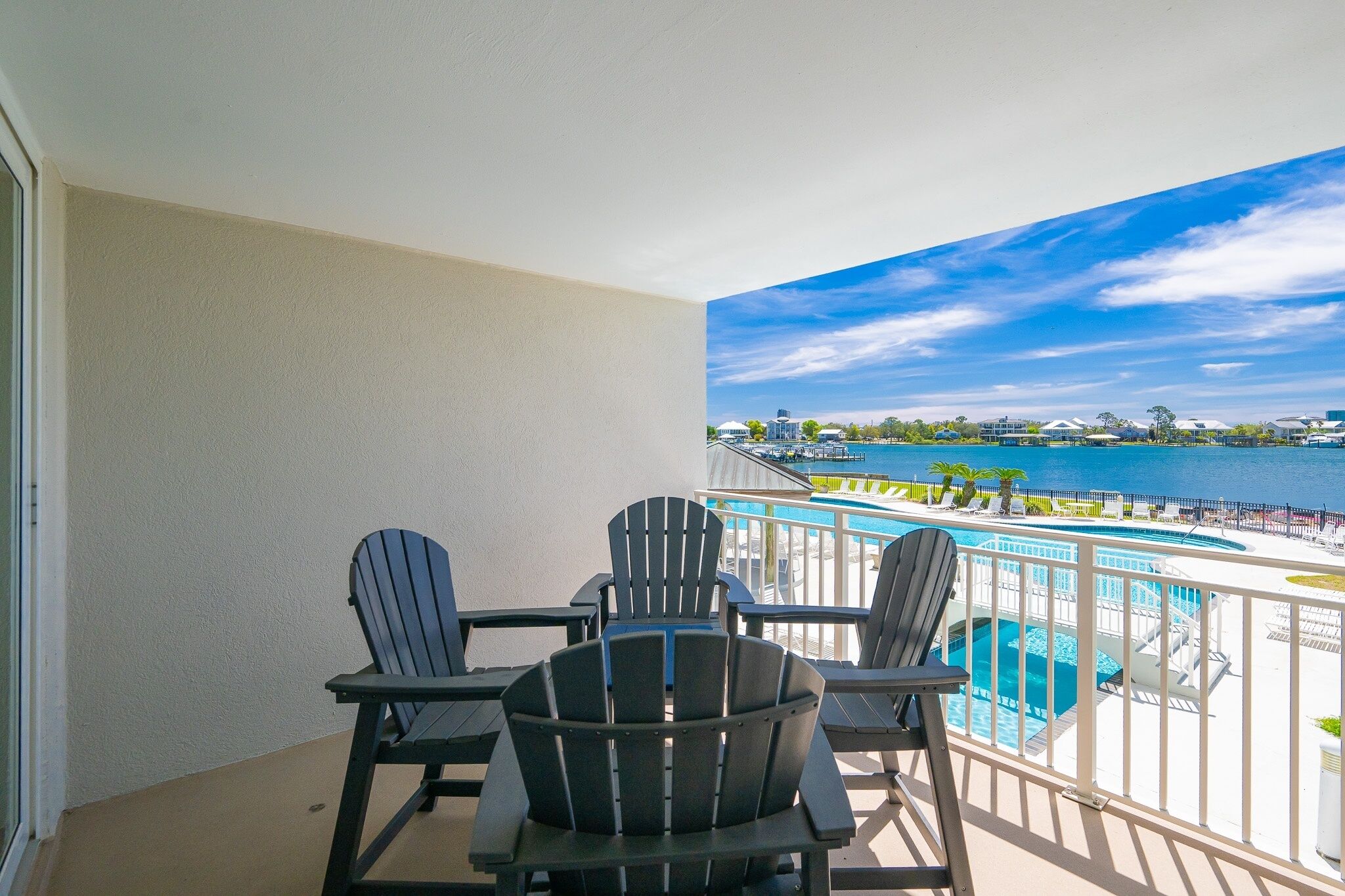 A balcony with three Adirondack chairs, a pool, and a view of a body of water and distant buildings.