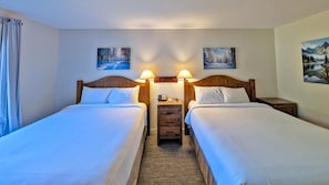 Habitación de hotel con dos camas, cabecera de madera, mesita de noche con lámpara y dos cuadros colgados en la pared.