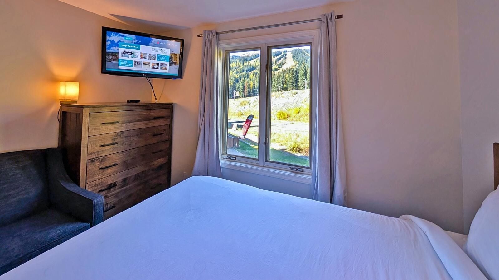 Ein Hotelzimmer mit einem Bett, einem an der Wand montierten Fernseher, einer Holzkombination und einem Fenster mit Blick auf eine gebirgige Landschaft.